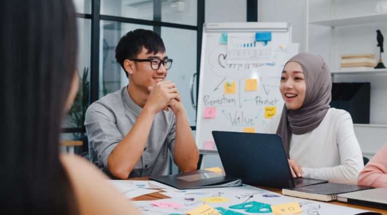 Gambar ini menunjukkan dua profesional yang sedang berdiskusi di ruang rapat, dengan papan tulis dan laptop sebagai alat bantu kerja mereka.