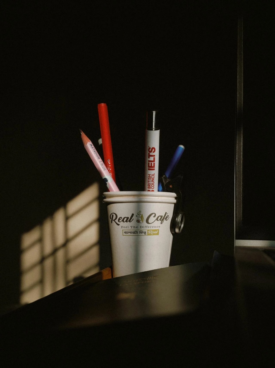 Sebuah cangkir kertas di atas meja berisi berbagai alat tulis, dengan spidol berlogo 'IELTS' terlihat jelas di bawah cahaya dari jendela.