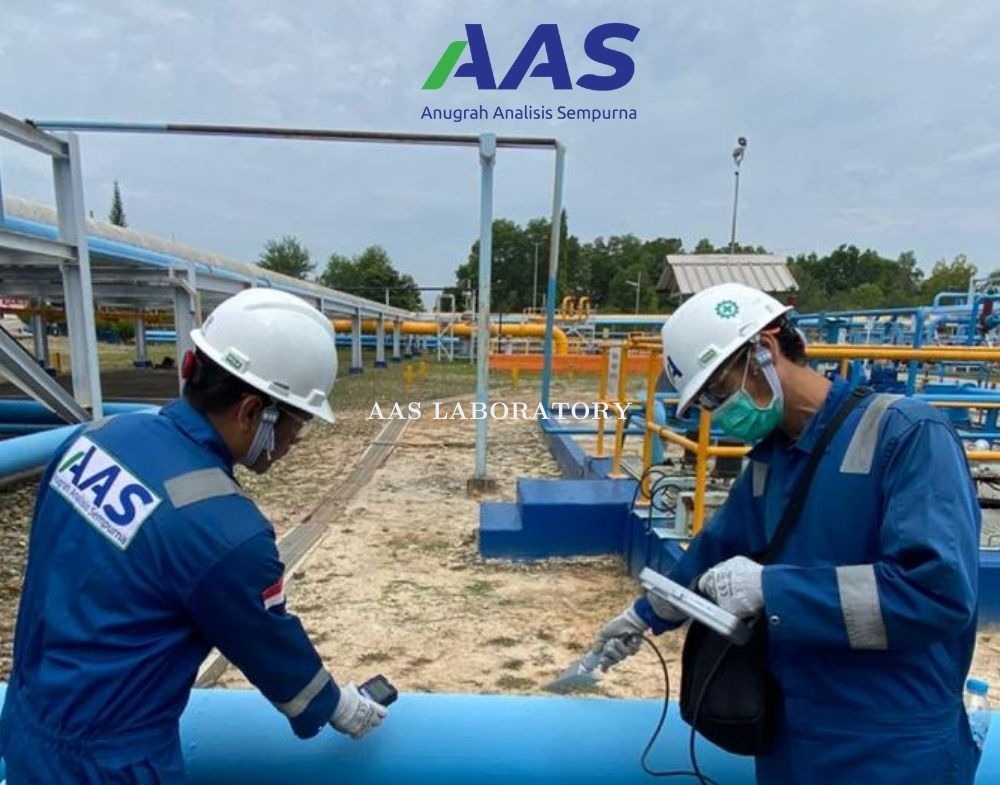 Dua teknisi AAS Laboratory dengan seragam biru dan helm putih melakukan pengambilan sampel atau inspeksi di fasilitas industri